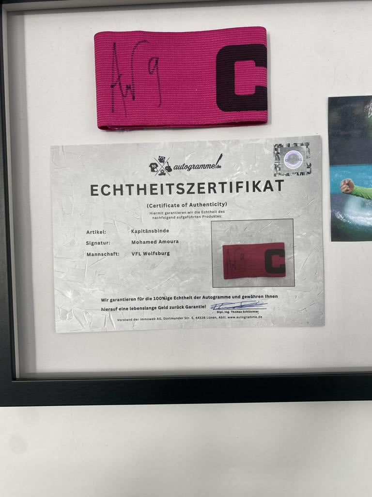 Brazalete de capitán firmado por Mohamed Amoura en un marco de Wolfsburg, certificado de autenticidad.