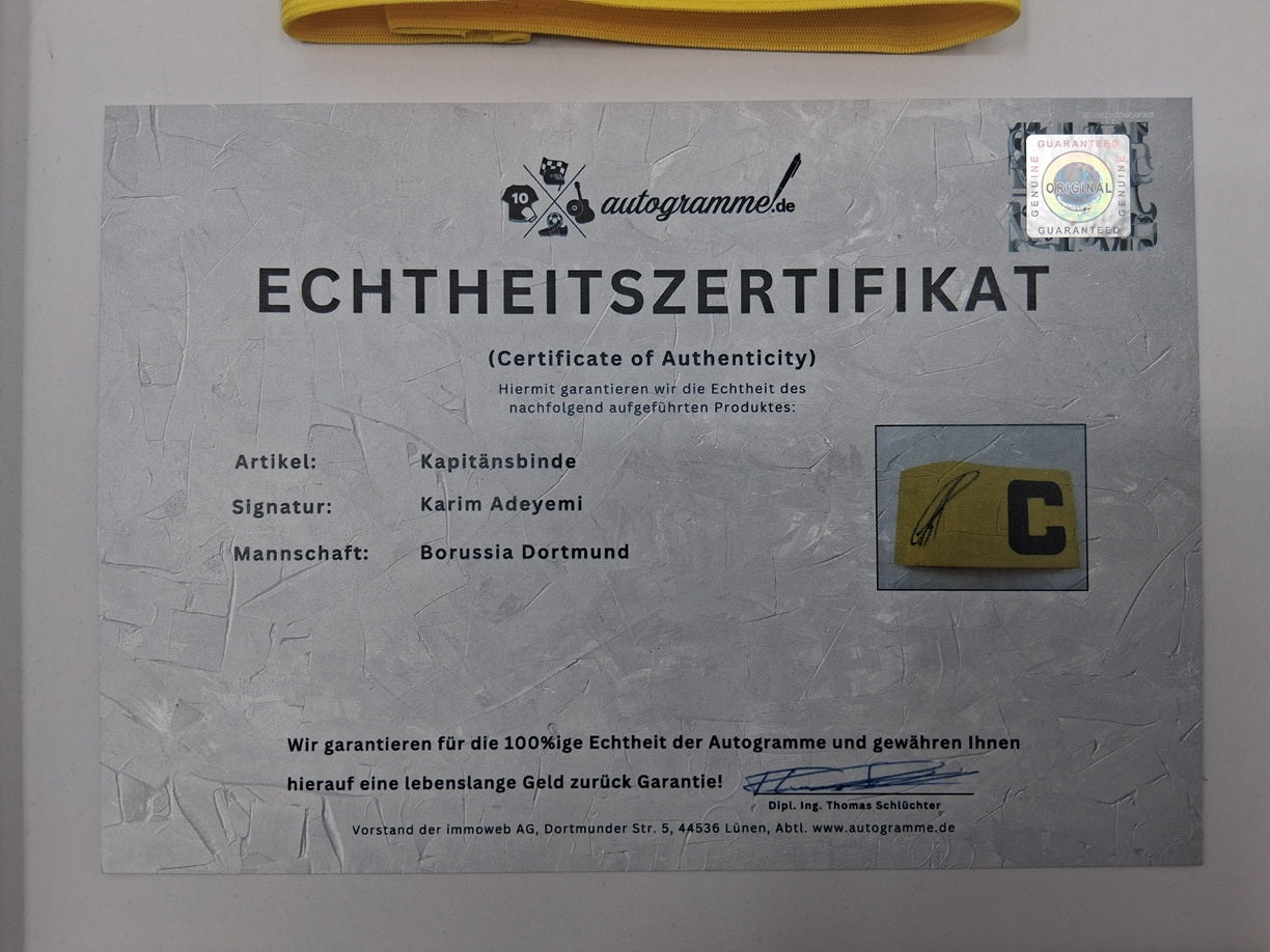 Brazalete de capitán firmado por Karim Adeyemi en un marco con certificado de autenticidad del Borussia Dortmund.